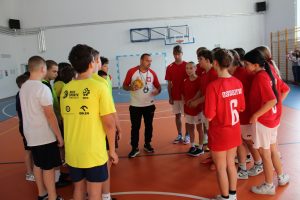 Trener w biało-czerwonej koszulce stoi na środku sali gimnastycznej i mówi do grupy młodzieży w żółtych i czerwonych koszulkach sportowych, zgromadzonych wokół niego.