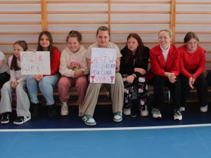 Uczennice siedzą na ławce w sali gimnastycznej, trzymają plakaty z hasłami dopingującymi: „Polska góra klasa 6” i „Moc jest w nas”.