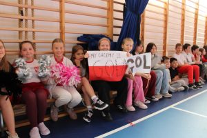 Grupa dzieci siedzi na ławce w sali gimnastycznej. Kilkoro z nich trzyma transparenty z napisami „My chcemy gol” i „Gol”, inne mają pompony w dłoniach.