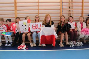 Dzieci siedzą w szeregu na ławce w sali gimnastycznej. Trzymają biało-czerwone plakaty i flagi z napisami „Gol” oraz rysunkami piłek.