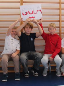 Trzech chłopców siedzi na ławce w sali gimnastycznej. Środkowy trzyma nad głową plakat z napisem „GOLA” w biało-czerwonych barwach.