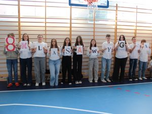 Grupa uczniów stoi w szeregu w sali gimnastycznej, każdy trzyma kartkę z literą. Razem tworzą napis „8 klasa wygrywa”.