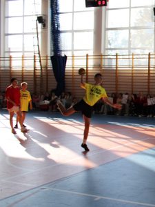 Chłopak w żółtej koszulce sportowej wyskakuje z piłką w ręku, przygotowując się do rzutu, podczas meczu na sali gimnastycznej.