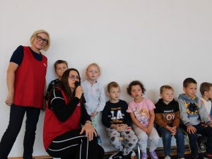 Grupa małych dzieci siedzi na ławce przy ścianie sali gimnastycznej. Obok nich stoją dwie nauczycielki w czerwonych kamizelkach, jedna trzyma mikrofon.
