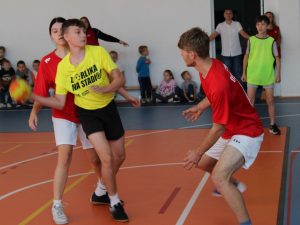 Chłopak w żółtej koszulce z piłką w ręku próbuje minąć dwóch zawodników w czerwonych koszulkach podczas meczu.