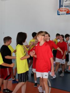 Zawodnicy w czerwonych i żółtych koszulkach podają sobie dłonie po meczu na sali gimnastycznej.