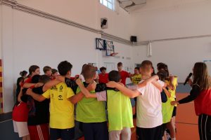 Grupa uczniów w czerwonych i żółtych koszulkach stoi w kręgu na sali gimnastycznej, obejmując się nawzajem ramionami.