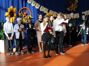 Grupa uczniów występuje podczas szkolnego apelu z okazji Dnia Edukacji Narodowej. Dzieci stoją na tle niebieskiej kotary z napisem „Dzień Edukacji Narodowej” oraz ozdobami z kwiatów i pomalowanych kół rowerowych. Występujący mają różne rekwizyty i stroje symbolizujące różne zawody i role szkolne.