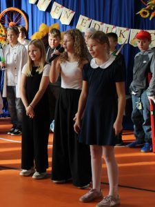 Trzy uczennice stoją na scenie w sali gimnastycznej, a jedna z nich trzyma mikrofon i mówi. W tle widać innych uczniów w kostiumach i napis „Dzień Edukacji Narodowej” zawieszony na niebieskiej kurtynie z ozdobami w kształcie kwiatów i kół rowerowych.