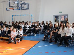 Uczniowie i nauczyciele siedzą na widowni w sali gimnastycznej podczas apelu z okazji Dnia Edukacji Narodowej. Dzieci i młodzież mają odświętne stroje, a w tle widać kosz do koszykówki i bramkę sportową.