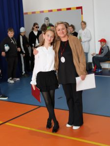 Uczennica i nauczycielka pozują do zdjęcia na sali gimnastycznej podczas apelu z okazji Dnia Edukacji Narodowej. Dziewczynka trzyma czerwoną teczkę, nauczycielka arkusz papieru. W tle widać innych uczniów w przebraniach i ozdoby na niebieskiej kotarze.
