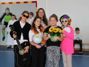 Grupa uczennic i nauczycielka pozują razem na tle szkolnej ściany. Dzieci mają kolorowe przebrania i rekwizyty: gitara, globus, konewka z kwiatami i paleta malarska. W tle widoczni są inni uczniowie w białych strojach.