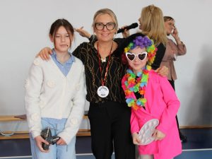 Dwie uczennice i nauczycielka stoją razem w sali gimnastycznej. Jedna dziewczynka trzyma metalowy imadło, druga jest przebrana w różowy żakiet, tęczową perukę i okulary w kształcie serc. W tle widać osoby prowadzące apel.
