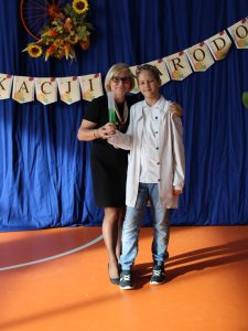 Nauczycielka i uczeń pozują do zdjęcia na tle niebieskiej kotary z napisem „Dzień Edukacji Narodowej”. Kobieta ubrana na czarno trzyma w ręku cylinder z zielonym płynem, a chłopiec w białym fartuchu laboratoryjnym stoi obok niej, uśmiechając się do aparatu.