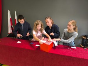 Czworo uczniów przygotowuje urnę wyborczą przed rozpoczęciem głosowania. Dzieci siedzą za stołem przykrytym czerwonym obrusem i naklejają oznaczenia na czerwoną skrzynkę wyborczą. W tle widoczne są biało-czerwone flagi.