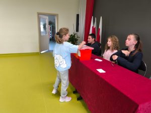 Uczennica wrzuca kartę do czerwonej urny wyborczej. Za stołem siedzą członkowie komisji uczniowskiej. W tle widoczne są flagi narodowe i dekoracja sali.