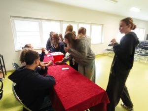Grupa uczniów stoi przy stole przykrytym czerwonym obrusem. Uczniowie podchodzą, aby oddać głos w wyborach do samorządu uczniowskiego. W tle widać rzędy krzeseł i jasne okna.