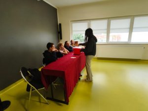 Uczennica oddaje głos, wrzucając kartę do czerwonej urny wyborczej. Za stołem siedzą członkowie komisji wyborczej obserwujący przebieg głosowania.