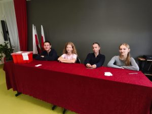 Czworo uczniów siedzi za stołem komisji wyborczej. Na stole znajduje się czerwona urna z kartami do głosowania. W tle widać flagi narodowe i dekorację sali.