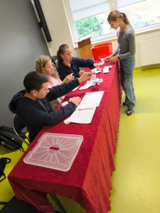 Członkowie komisji wyborczej liczą oddane głosy. Na stole leżą karty wyborcze i arkusz do zapisywania wyników. Uczniowie pracują wspólnie, zachowując skupienie.