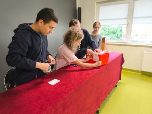 Uczniowie przygotowują urnę wyborczą przed rozpoczęciem głosowania. Naklejają oznaczenia na czerwoną skrzynkę stojącą na stole przykrytym czerwonym obrusem.