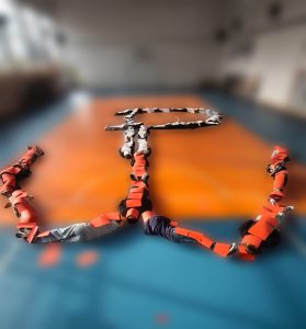 Uczniowie leżą na sali gimnastycznej, tworząc kształt kotwicy Polski Walczącej z użyciem czerwonych kartek.