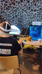 Młody chłopak siedzi przy biurku, gra na komputerze w symulator jazdy samochodem, korzystając z zestawu VR i kierownicy. Na koszulce ma napis „Technik informatyk e-sportowa”. Obok monitora stoi komputer z podświetleniem w kolorze niebieskim i mały żółty robot-zabawka.
