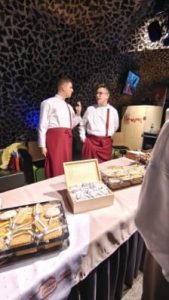 Dwóch młodych mężczyzn w białych koszulach i bordowych fartuchach stoi za stołem nakrytym obrusem. Na stole leżą tace z zapakowanymi przekąskami oraz otwarte pudełko z ciastkami owiniętymi w folię. Mężczyźni rozmawiają ze sobą, a w tle widać ciemną ścianę o geometrycznym wzorze i oświetlenie sceniczne.