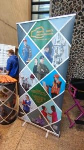Baner promocyjny szkoły pokazujący różne kierunki kształcenia, w tym mundurowe, informatyczne i gastronomiczne. Baner ma układ złożony z kilku zdjęć uczniów w różnych strojach zawodowych. Stoi przy ścianie o geometrycznym wzorze, obok stolika z przekąskami i osoby w niebieskiej bluzie.