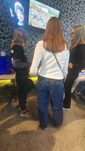 Trzy młode osoby stoją przed stanowiskiem komputerowym z dużym monitorem wyświetlającym animowaną postać i widok miasta. Jedna z nich ma białą bluzę, druga czarną, a trzecia jasne włosy i okulary. W tle widać komputer z niebieskim podświetleniem i charakterystyczną ścianę o geometrycznym wzorze.