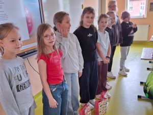 Grupa dziewczynek stoi w szeregu w klasie przy tablicy multimedialnej. Przed każdą z nich leży kolorowa torba upominkowa przygotowana z okazji Dnia Chłopaka.