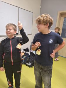 Dwóch chłopców stoi obok siebie i trzyma na sznurkach jabłka z wygryzionymi kawałkami. Jeden z chłopców ma na sobie czarną bluzę z czerwonym zamkiem, drugi granatową koszulkę. W tle widać plecaki i krzesła.