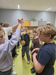 Grupa dzieci w klasie uczestniczy w zabawie z jabłkami na sznurkach. Jedna osoba trzyma jabłko zawieszone na sznurku, a inne dzieci patrzą i czekają na swoją kolej. W tle widać szafki i krzesła.