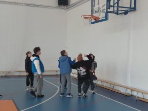 Grupa chłopców gra w koszykówkę w sali gimnastycznej. Widać moment pod koszem, gdy kilku z nich patrzy w górę na obręcz na której wisi karteczka z zadaniem.