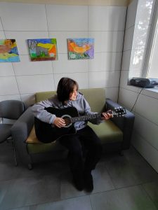 Chłopiec w szarej bluzie siedzi na zielonej sofie i gra na czarnej gitarze akustycznej. W tle na ścianie wiszą kolorowe dziecięce rysunki zwierząt.