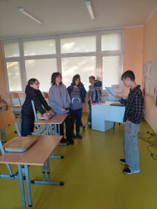 Kilkoro uczniów stoi i siedzi w klasie przy ławkach. Jeden chłopiec z przodu czyta coś z kartki, pozostali uważnie go słuchają.