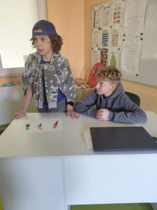 Dwóch chłopców stoi za biurkiem nauczycielskim. Jeden ma czapkę z daszkiem i bluzę przewieszoną przez ramiona, drugi opiera się o blat z dłońmi splecionymi. Na biurku leżą trzy klucze na brelokach i laptop.