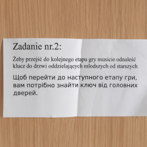 Kartka papieru przyklejona do drewnianych drzwi. Na kartce wydrukowany jest tekst w dwóch językach: po polsku i po ukraińsku. Nagłówek brzmi „Zadanie nr 2”, a treść informuje, że aby przejść do kolejnego etapu gry, należy odnaleźć klucz do drzwi oddzielających młodszych od starszych.