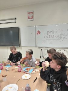 Grupa chłopców siedzi przy stole w klasie i je pizzę. Na tablicy w tle napis „Dzień Chłopaka – wszystkiego najlepszego!!! 6/7”. Na stole stoją napoje, talerze i przekąski.