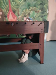 Chłopiec siedzi skulony pod stołem do piłkarzyków, przy ścianie z kolorowym, zwierzęcym muralem. Widać jego białe skarpetki i przytulone do siebie nogi.