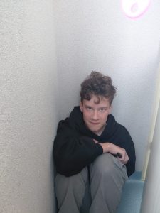 Chłopiec w czarnej bluzie siedzi skulony w wąskim rogu przy ścianie, patrzy prosto w obiektyw.