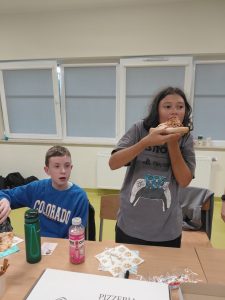Dwóch chłopców przy stole w klasie. Jeden w niebieskiej bluzie siedzi i patrzy w bok, drugi w szarej koszulce z nadrukiem trzyma dużą porcję pizzy i je. Na stole napoje, karton z pizzerii i przekąski.