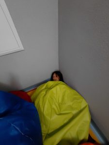 Chłopiec owinięty dużym zielonym workiem-pufą siedzi w rogu sali z szarymi ścianami. Obok widoczne są kolorowe pufy.