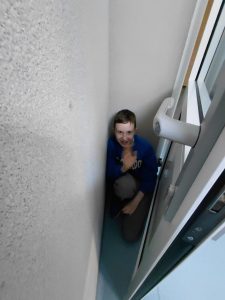 Chłopiec w niebieskiej bluzie siedzi skulony w wąskim kącie między ścianą a drzwiami, uśmiecha się i unosi rękę w geście machania.