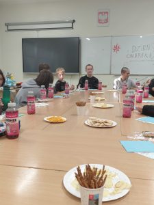 Uczniowie siedzą przy dużym stole w klasie. Na stole ustawione są talerze z przekąskami, precelki i napoje. W tle na tablicy napis „Dzień Chłopaka – wszystkiego najlepszego”.