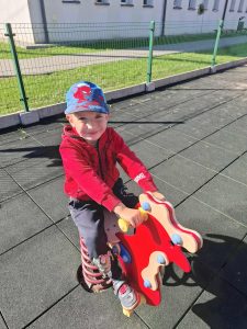 Uczeń oddziału przedszkolnego siedzi na sprężynowym bujaku w kształcie zwierzątka na szkolnym placu zabaw. Uśmiecha się do aparatu, a w tle widać budynek szkoły i ogrodzenie.
