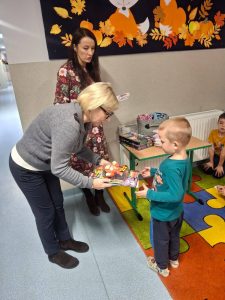 Pani dyrektor wręcza nagrody chłopcu na szkolnym korytarzu. Obok stoi nauczycielka, a na stoliku znajdują się upominki. W tle dzieci siedzą na kolorowym dywanie.