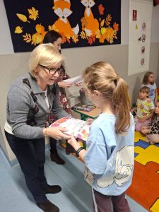 Pani dyrektor wręcza dyplom uczennicy na szkolnym korytarzu. Za nimi znajduje się jesienna dekoracja z lisami i liśćmi oraz dzieci siedzące na dywanie.