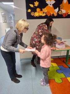 Na szkolnym korytarzu pani dyrektor wręcza dyplom dziewczynce. W tle widoczna nauczycielka przy stole z nagrodami oraz jesienna dekoracja z lisami i liśćmi.