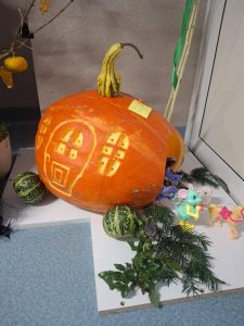 Duża dynia ozdobiona na domek z wyciętymi oknami i drzwiami. Przy dyni stoją małe zielone tykwy, gałązki iglaste oraz zabawkowe zwierzątka, w tym myszka i konik.